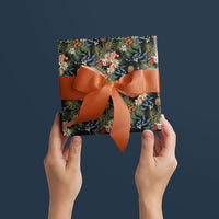 Hands holding a wrapped gift box with dark floral wrapping paper and burnt orange ribbon bow against blue background. Illustrated by Lisel Ashlock.