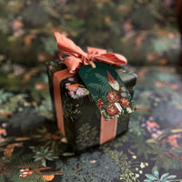 A decorative gift tag with gnome and mushroom illustrations on dark green cardstock tied with a coral ribbon. Illustrated by Lisel Ashlock.