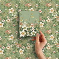 A hand holds a green thank you greeting card with white flowers against a coordinating floral pattern background. Illustrated by Lisel Ashlock.