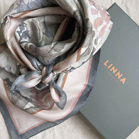 Folded Angelite silk cotton scarf in cream, gray, and mauve tones with botanical leaf motifs beside LINNA packaging. Illustrated by Lisel Ashlock.