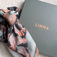 A folded silk cotton scarf with floral botanical print in rust, pink, and cream tones next to its gray LINNA California box. Illustrated by Lisel Ashlock.
