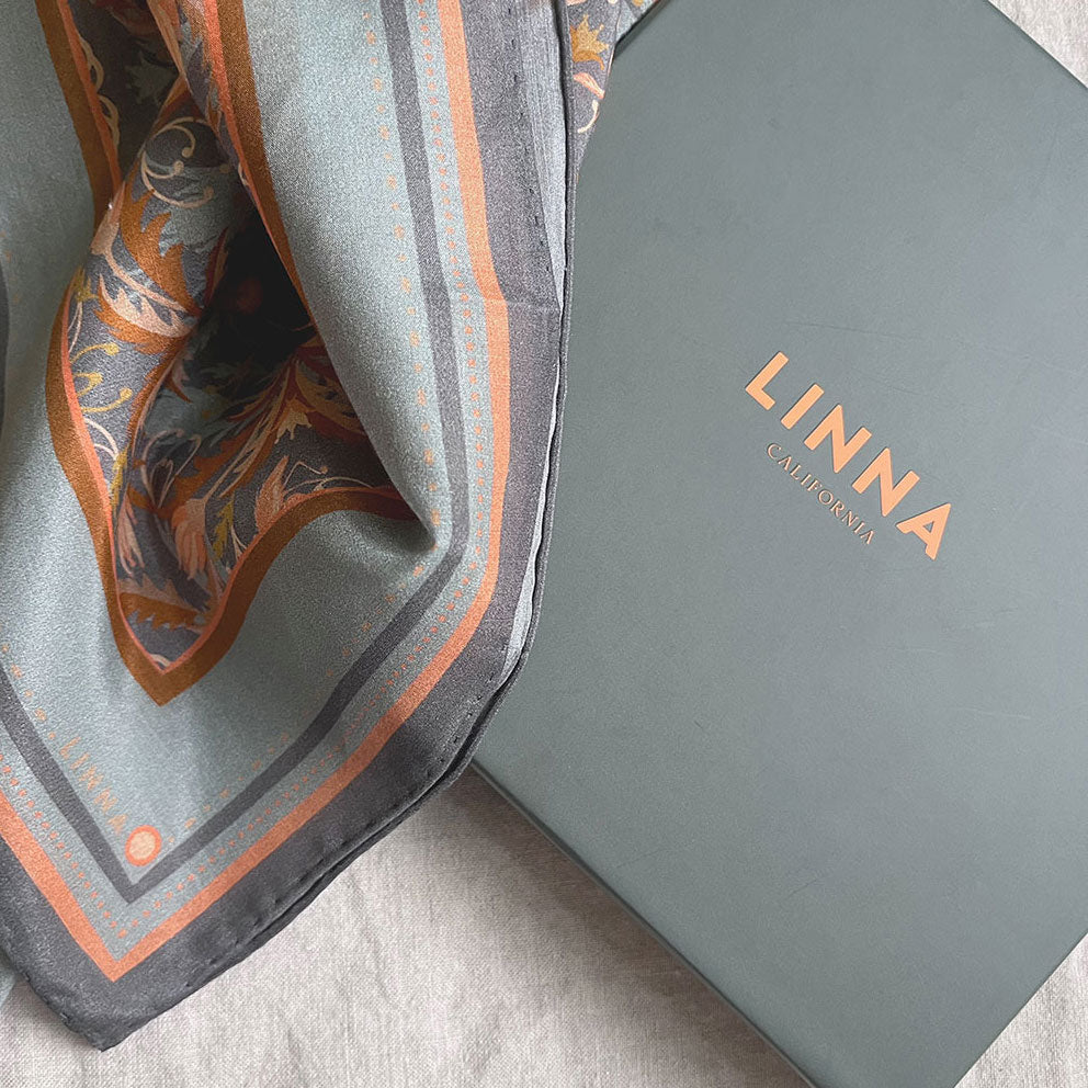 A folded apricot filigree silk cotton scarf next to its Linna California branded box on a neutral surface.
