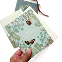 A hand holding an engagement greeting card with botanical illustrations and two butterflies, with a matching card behind it. Illustrated by Lisel Ashlock.