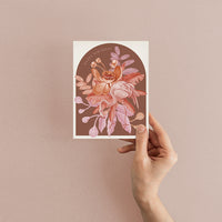 Hand holding a birthday greeting card with illustrated flowers and a bee on brown background by Lisel Ashlock.