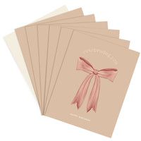 Stack of greeting cards with beige covers featuring a pink ribbon bow and text reading "celebrating you" and "happy birthday." Illustrated by Lisel Ashlock.