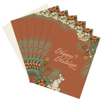 A fanned stack of Happy Holidays greeting cards featuring illustrated holly berries, pine branches, pinecones, and a white hare on a rust background. Illustrated by Lisel Ashlock.