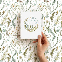 Hand holding a blank greeting card featuring botanical illustrations of wildflowers and herbs in green and gold tones. Illustrated by Lisel Ashlock.
