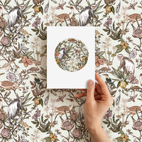 A hand holding a blank greeting card featuring a circular botanical illustration with monkeys and flowers against a patterned background. Illustrated by Lisel Ashlock.