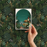 Hand holding a holiday greeting card featuring a whimsical gnome in an enchanted forest with "Peace & Love" text. Illustrated by Lisel Ashlock.