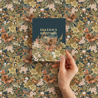 Hand holding a "Season's Greetings" card against a dark floral botanical pattern background. Illustrated by Lisel Ashlock.