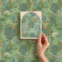 A hand holds a "Happy Birthday" greeting card featuring a woodland meadow illustration by Lisel Ashlock.