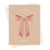 A beige greeting card with "celebrating you" text and a pink ribbon bow illustration, "Happy Birthday" below. Illustrated by Lisel Ashlock.