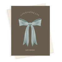 A brown greeting card featuring "celebrating you" text above an illustrated blue bow and "HAPPY BIRTHDAY" below. Illustrated by Lisel Ashlock.