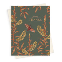 A greeting card with a dark green background featuring autumn botanical illustrations in gold and rust tones with a burgundy moth and "giving THANKS" text. Illustrated by Lisel Ashlock.