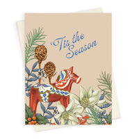 A festive greeting card featuring a red Dala horse surrounded by pine cones, white flowers, and berries. Illustrated by Lisel Ashlock.