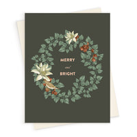A holiday greeting card with a dark green background featuring a wreath of green leaves, white flowers, and red berries, with gold text reading "MERRY and BRIGHT." Illustrated by Lisel Ashlock.