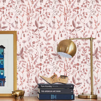 A wooden desk against pink floral wallpaper displays stacked art books, a wooden plane, and brass decorative objects under a brass lamp. Illustrated by Lisel Ashlock.