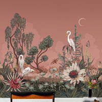 A coral pink mural wallpaper featuring white herons, flowering plants, trees, butterflies, and a crescent moon. Illustrated by Lisel Ashlock.