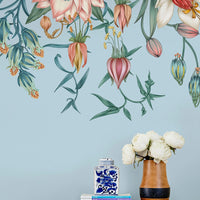 A light blue wall mural featuring hanging botanical illustrations in pink, coral, and green tones above a wooden shelf with books and white peonies. Illustrated by Lisel Ashlock.