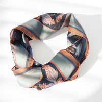 A folded silk cotton scarf in moonstone featuring botanical illustrations in copper, navy, and cream tones. Illustrated by Lisel Ashlock.