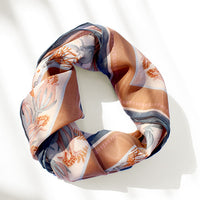 A rolled sunstone-colored silk cotton scarf with coral and navy botanical patterns cast in bright sunlight. Illustrated by Lisel Ashlock.